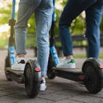 Two people on e-scooters. They each have a foot on an e-scooter and the other foot flat on the ground