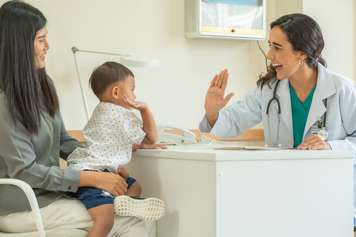 Health-care provider and child reaching to high-five across a desk between them. The child sits on a parent's lap.
