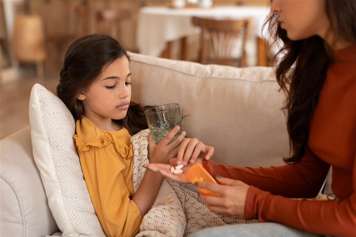 child sitting on couch holding a glass of water and taking a tablet from a woman's outstretched palm with her other hand