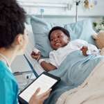 Child in a hospital bed with a pulse oximeter on his finger and an IV in his other hand. A nurse sits beside the bed