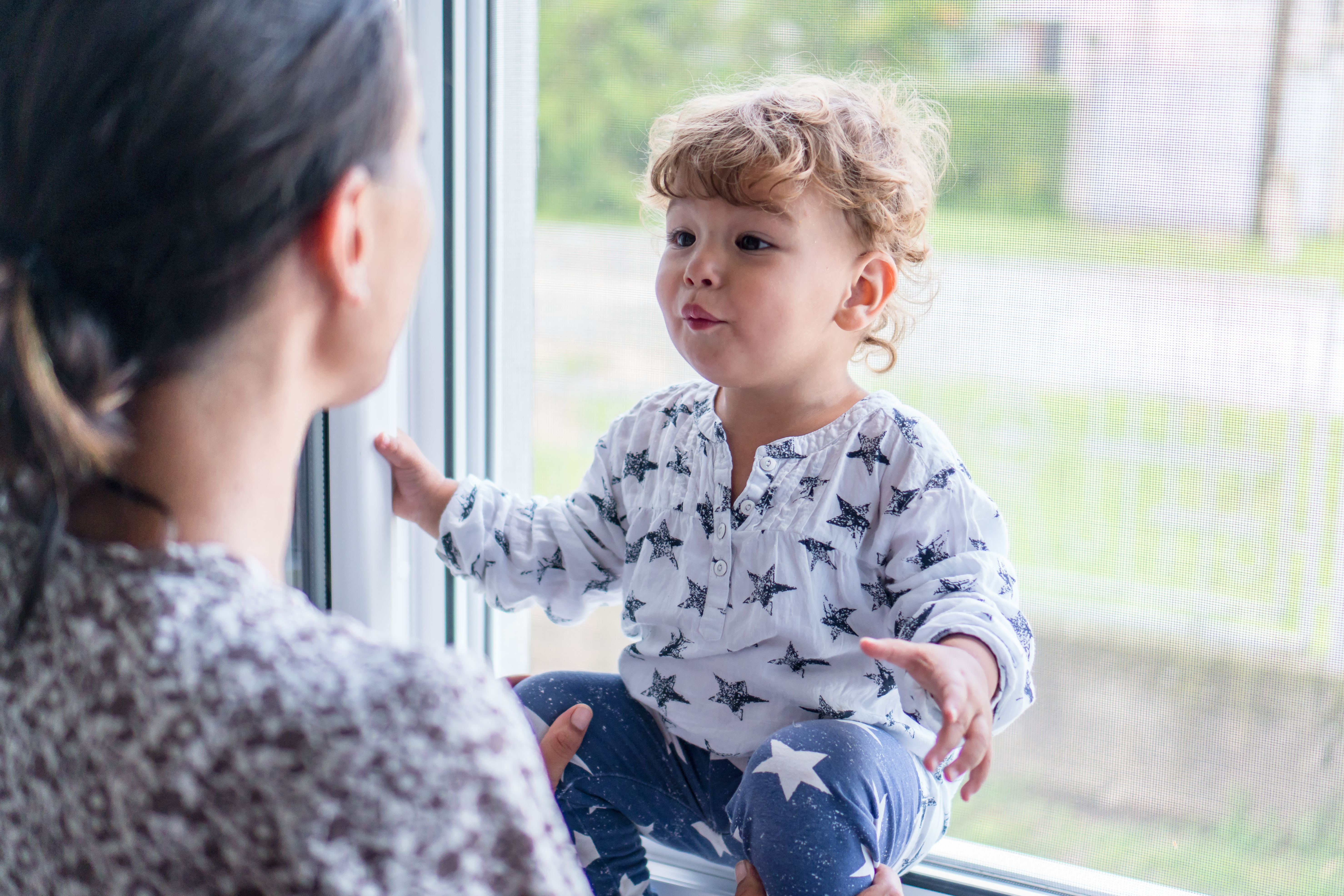 Toddler sitting on windowsill across from a woman who supports the toddler's legs with both hands