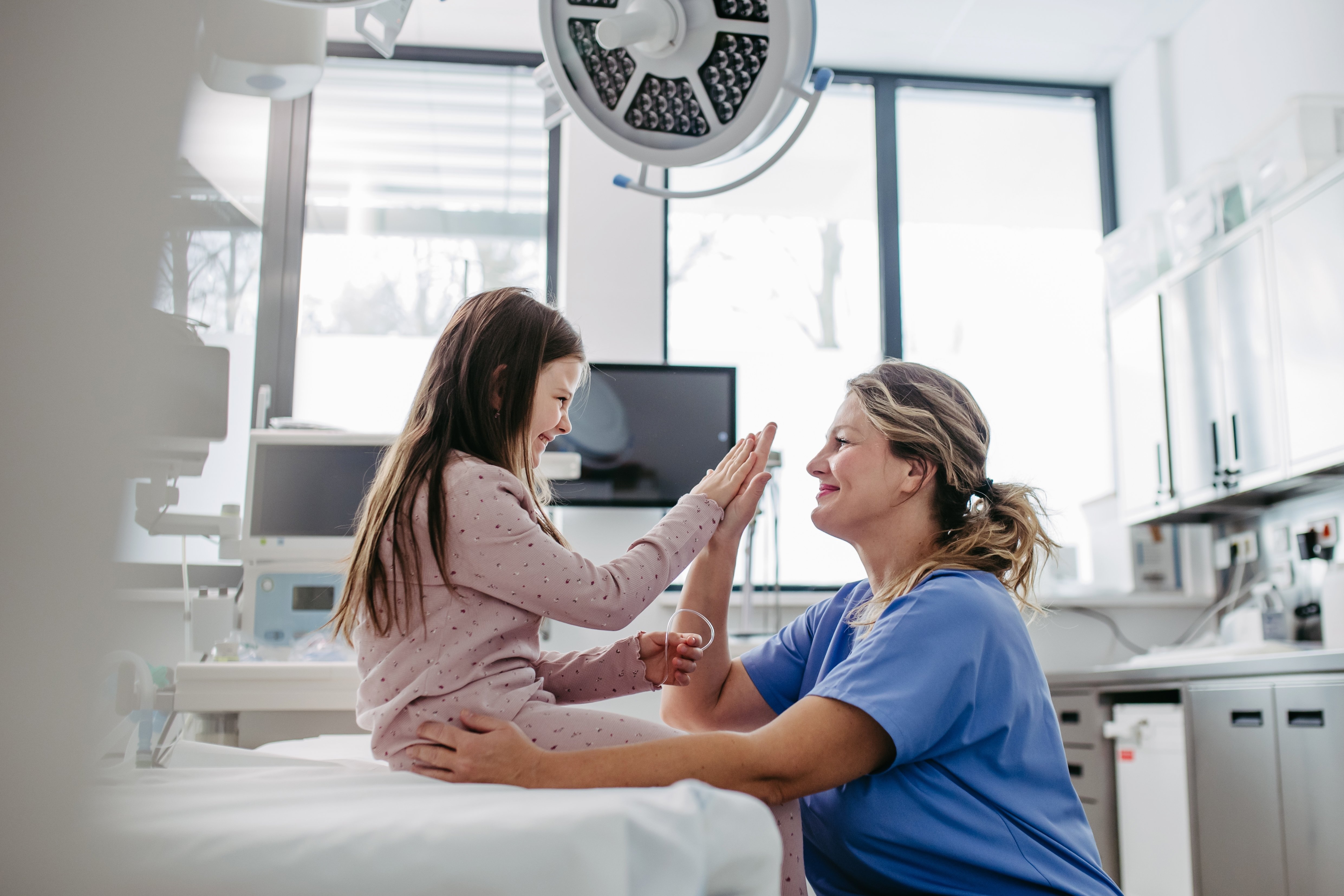 Child and health-care provider high-fiving. Child sits on hospital bed and health-care provider crouches in front of her