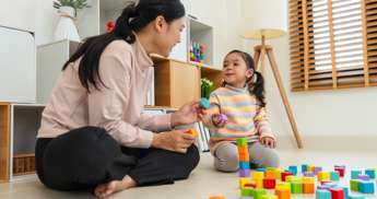 Woman and school-aged child sitting on a floor with colourful building blocks in front of them