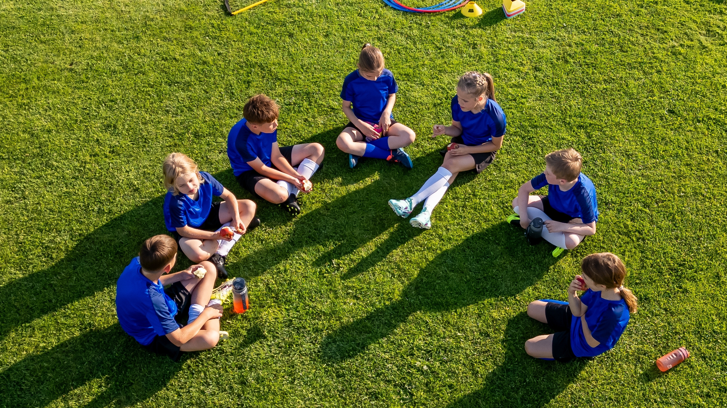 Overhead view of children in soccer uniforms sitting in a semi-circle on a grass field with sports equipment around them