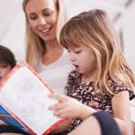 School-age girl sitting on a bed reading a book out loud. A woman and boy sit beside her